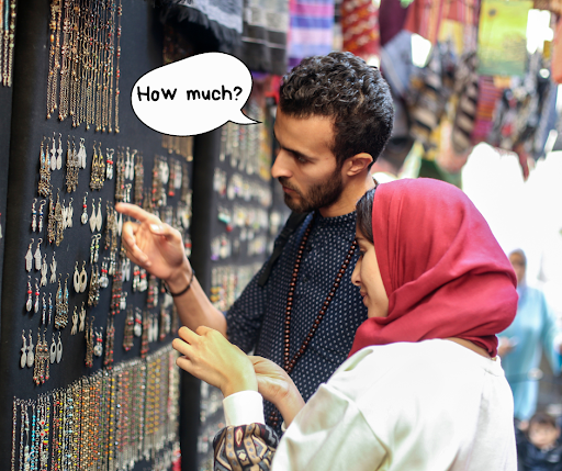 The image shows a couple at a market stall examining accessories. The man points at an item and asks, "How much?" while the woman, in a red hijab, looks on with interest. 